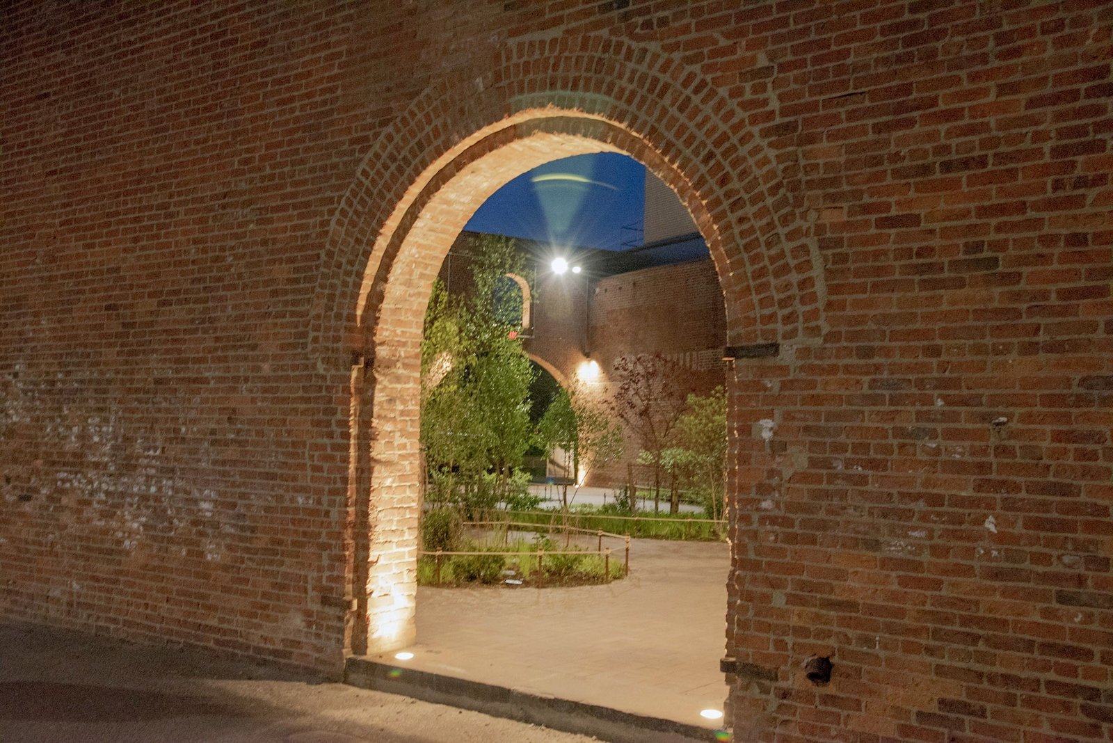 An illuminated brick arch frames a courtyard at night.